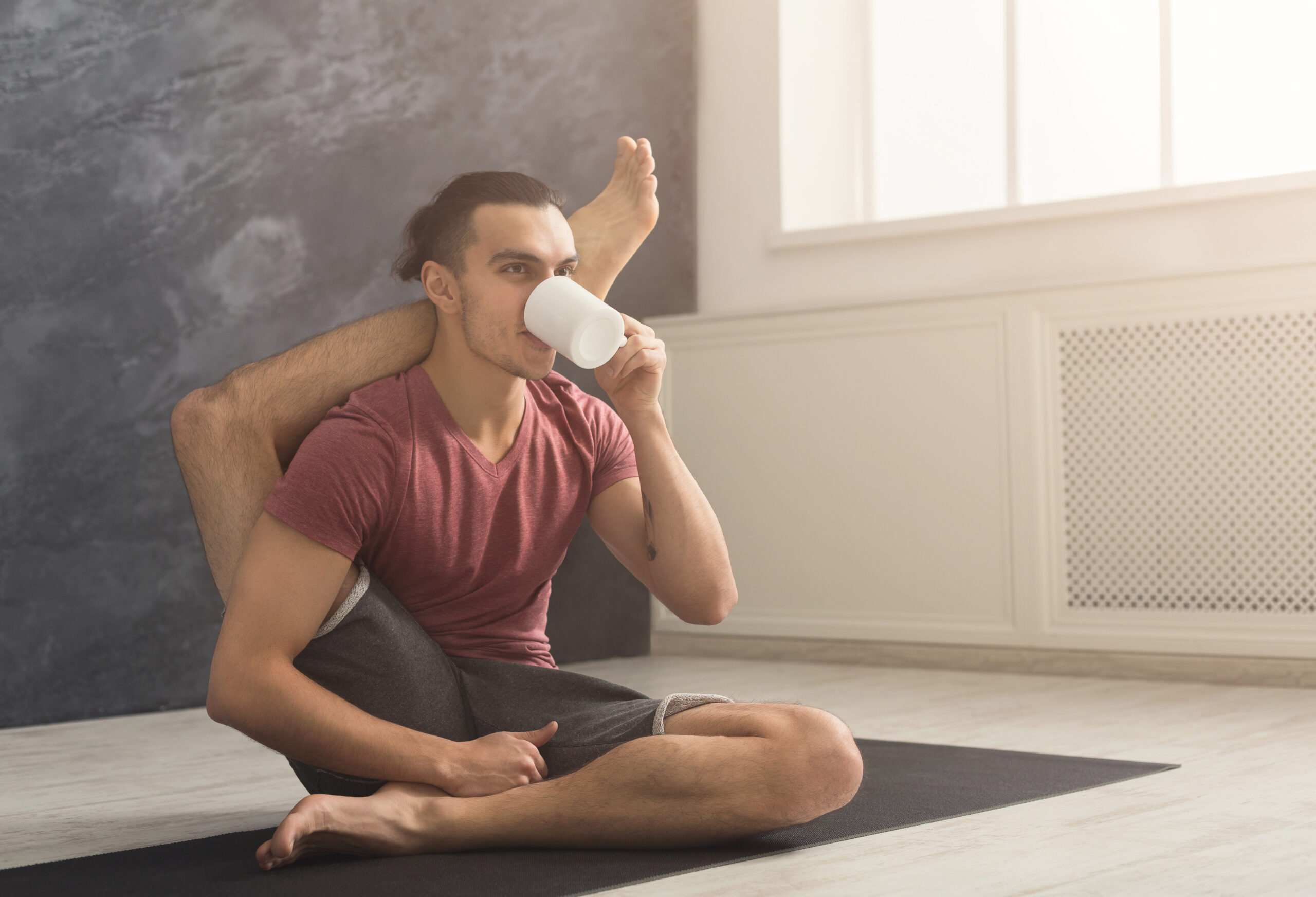 Young sporty man in flexible yoga pose drinking tea, sitting on mat at gym and practicing twisting exercise, copy space